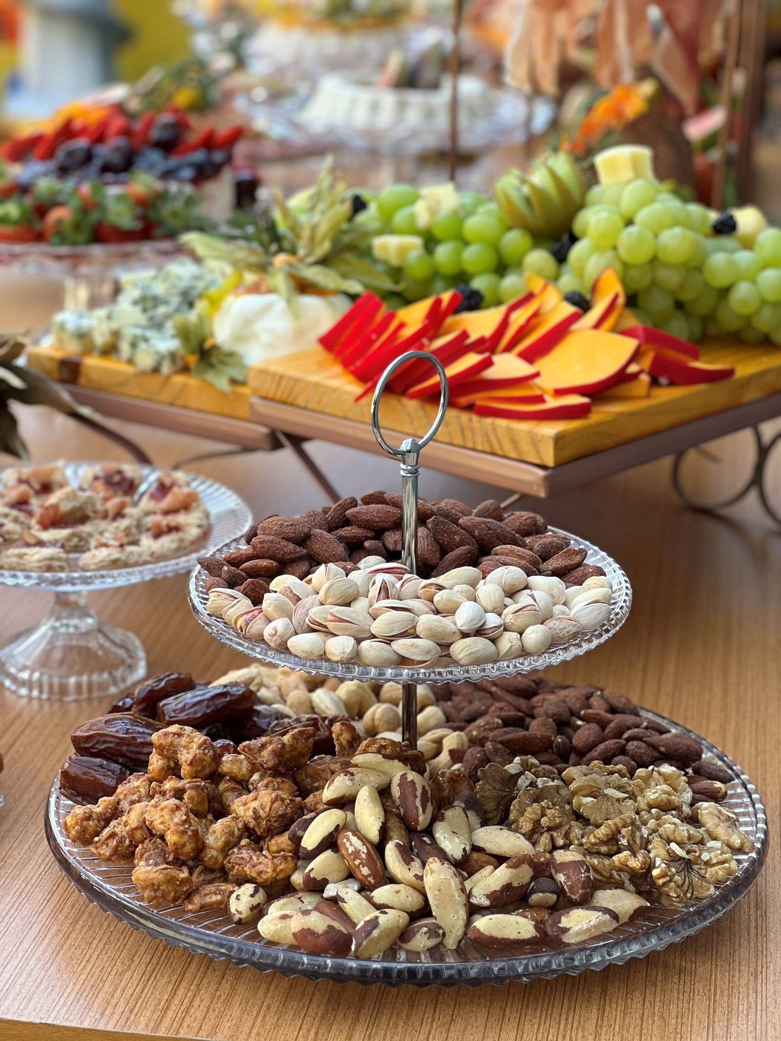 Prato de cristal em andares com amêndoas, pistaches, castanhas e tâmaras, com uvas, queijos e frutas ao fundo.