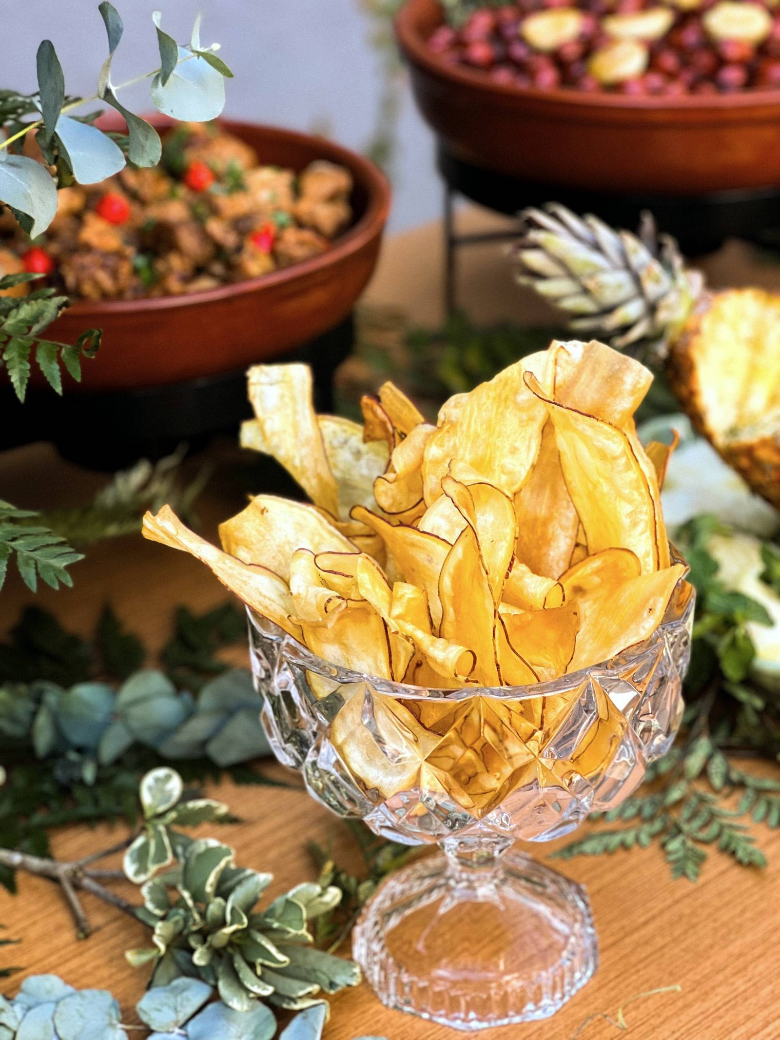 Taça de cristal lapidado cheia de chips dourados, sobre mesa de madeira com eucalipto.