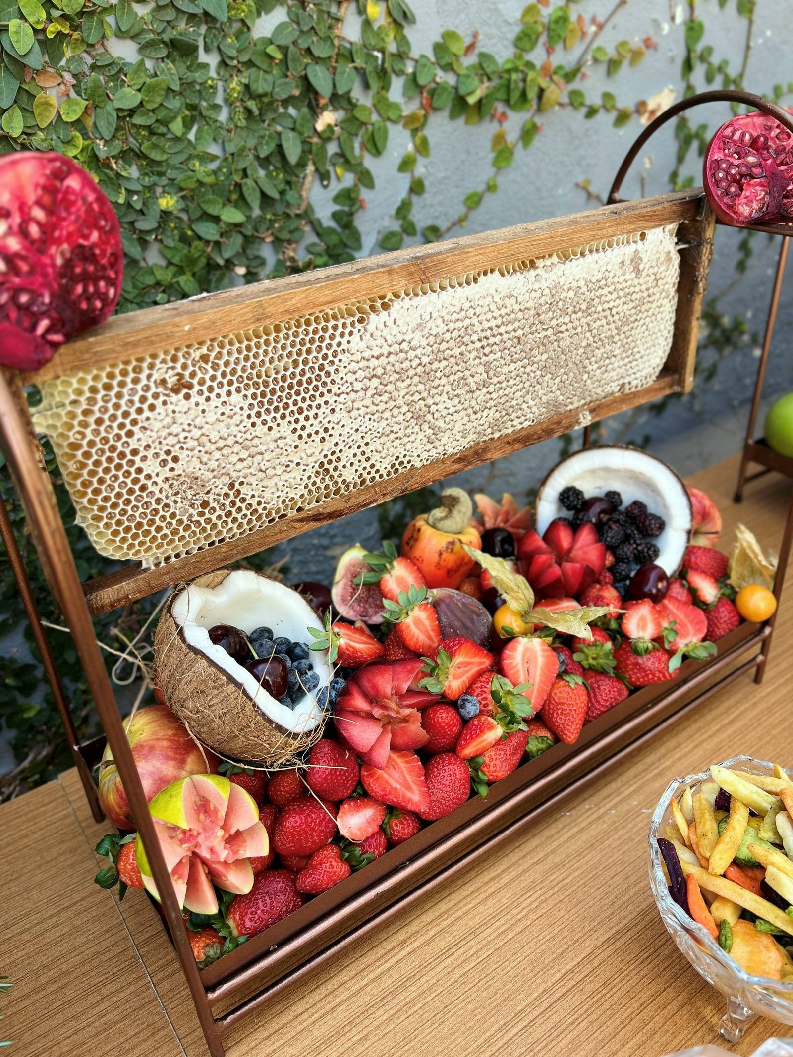 Suporte de ferro com favo de mel ao alto e bandeja de morangos, frutas vermelhas, coco e romãs.
