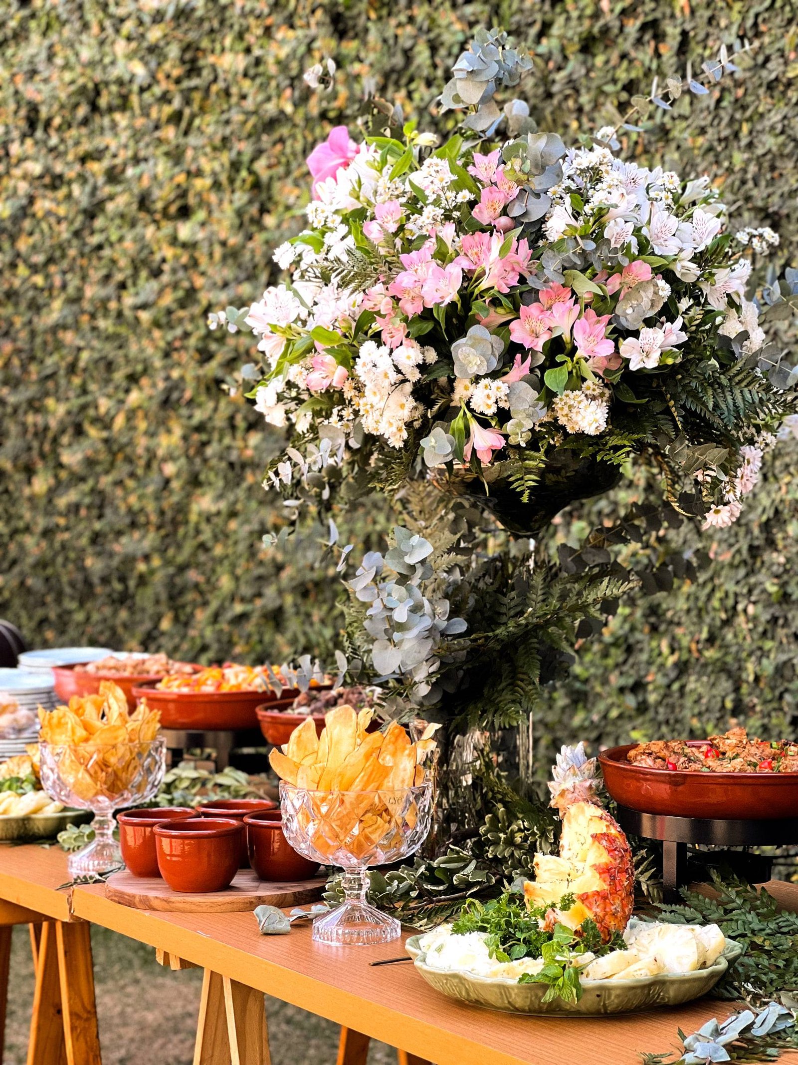 Mesa de buffet ao ar livre com grande arranjo de flores rosa e brancas, taças de cristal com chips e panelas de barro, diante de uma parede de folhagem.