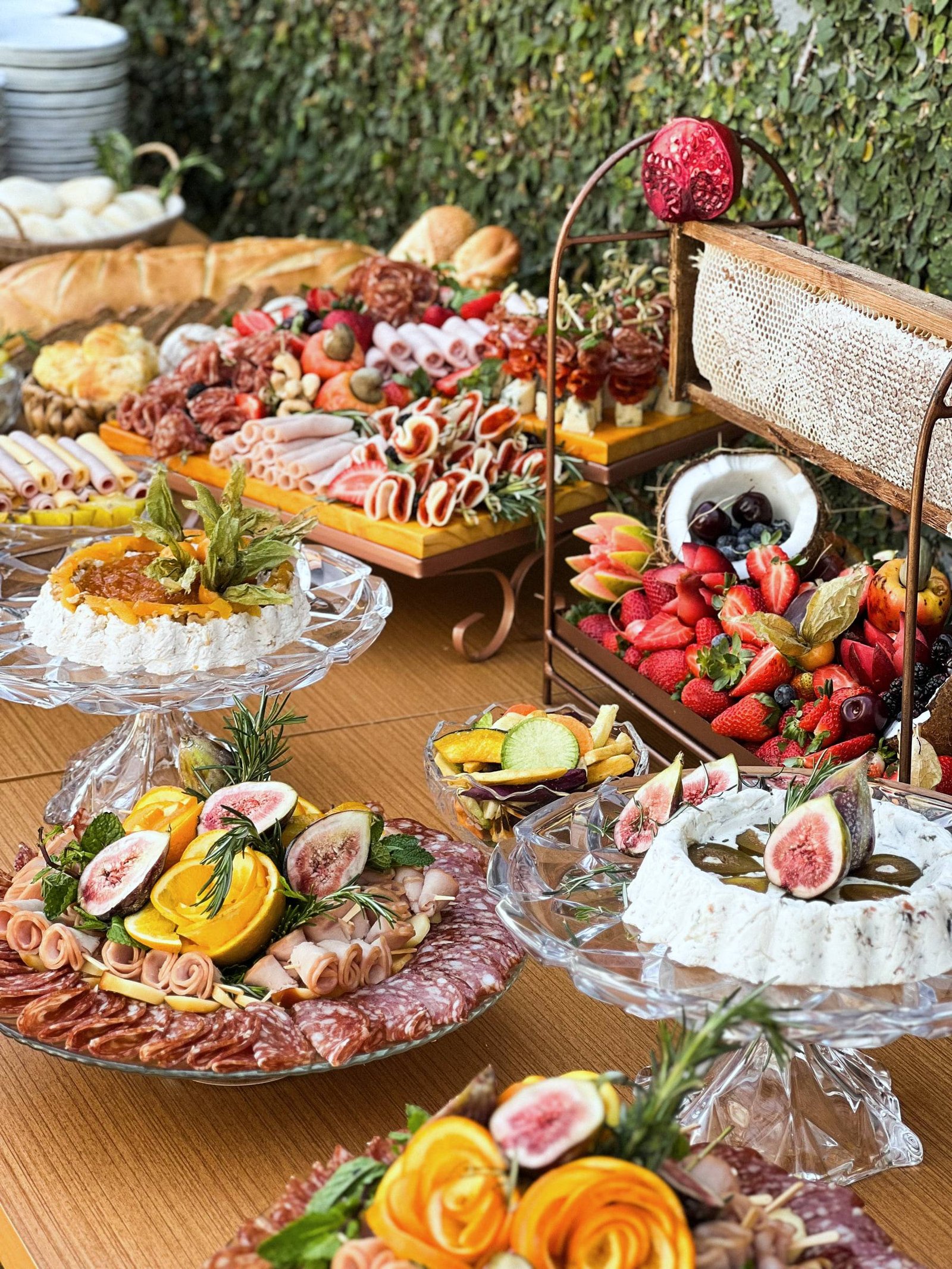 Mesa de buffet montada com frios, frutas, figos e frutas vermelhas em pratos de cristal e suportes de ferro, com favo de mel, diante de uma parede de folhagem.