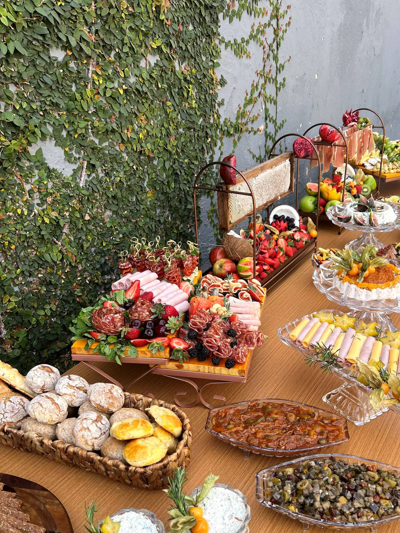 Mesa completa ao ar livre com cesta de pães, tábuas de frios, frutas, pastas e antepastos em travessas de cristal.