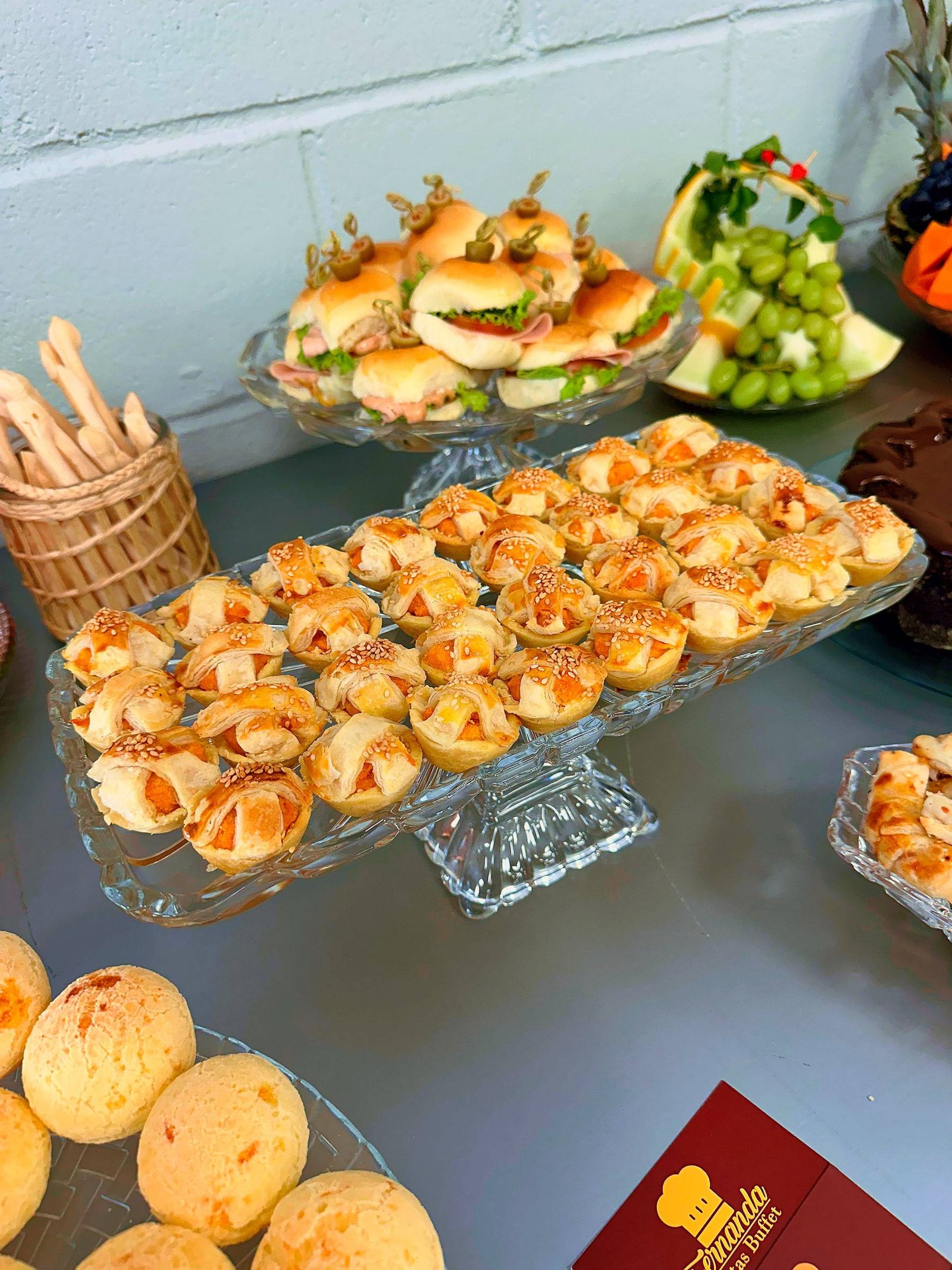 Travessa de cristal com salgados folhados, mini sanduíches ao fundo e pães de queijo em primeiro plano.