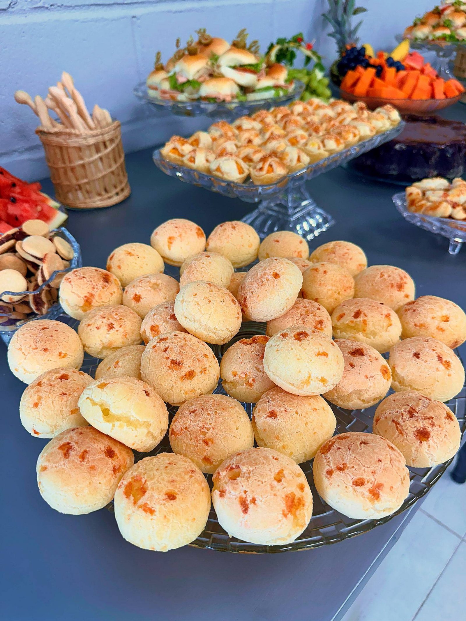 Travessa de cristal com pães de queijo, com mini sanduíches, folhados e frutas ao fundo.