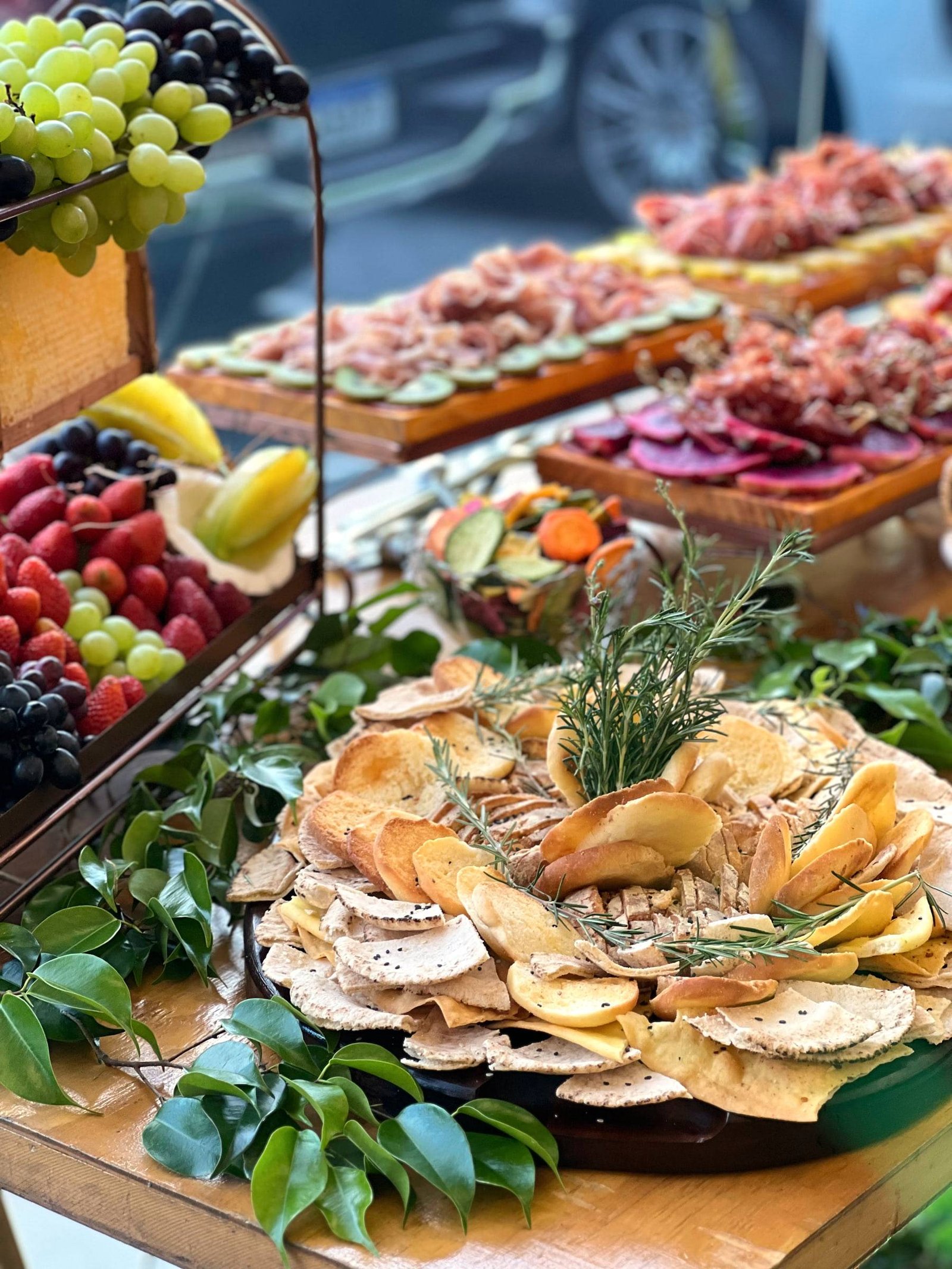 Travessa de torradas decorada com ramos de alecrim, com tábuas de frios e frutas ao fundo.